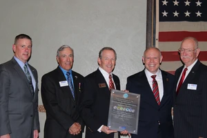 Five men in business suits stand shoulder to shoulder, smiling for a formal group photo. The two men in the center are holding a large framed award plaque, while a U.S. flag hangs in the background, giving the scene the feel of an official ceremony recognizing an organization or employer for its service and support.